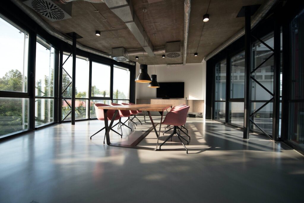 Modern office meeting room with large floor-to-ceiling windows providing natural light. Features a long wooden table surrounded by pink swivel chairs, industrial-style ceiling with exposed ducts, and hanging pendant lights above the table.